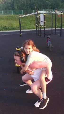A woman is giving a piggyback ride to a young girl. The woman is wearing a white t-shirt and pink shorts. She has headphones on. The girl is wearing a light-colored t-shirt. They are on a black rubber surface, likely a playground or outdoor gym. In the background, there are metal exercise structures. A bicycle is visible to the right. The sun is shining.
