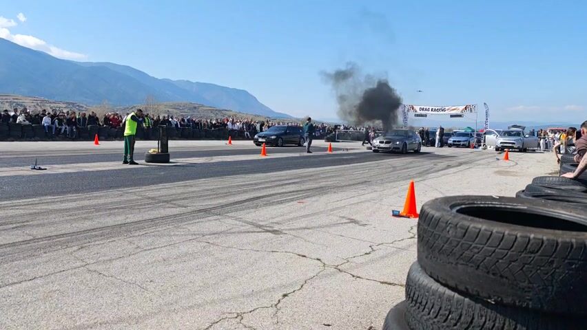 The scene shows two BMWs engaging in a drag race on an asphalt track. There is a line of spectators on both sides, and there are safety personnel and cones present. The car on the right is smoking heavily. The environment is a mountain valley under a blue sky with light clouds. The scene suggests a professional or organized drag racing event.