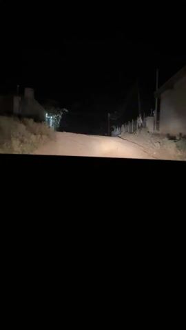 From a low angle at night, the view from inside a car looks out onto a dimly lit dirt road. A building with a partially blocked view sits to the left, illuminated by a street light. Further down the road, another structure is visible. To the right of the road, a simple fence runs along the property line, with tall wooden poles supporting wires above. The sky is dark and featureless. The dashboard of the car is black, creating a stark foreground. Music plays in the background.
