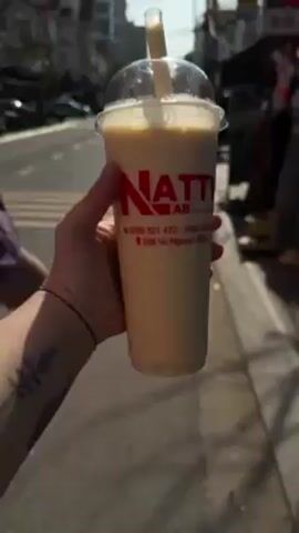 A person is holding a plastic cup with a drink with the "NATTY AB" logo on it. The drink is light brown and frothy. The background shows a street with pedestrians and buildings. The person is standing in front of a store with mannequins displaying clothing. The sky is clear.