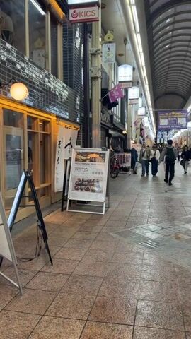 The video shows a busy, covered shopping arcade with a high, arched ceiling and overhead lights. Light-colored tiled flooring, some with patterns, extends down the arcade. On the left, an A-frame sign stands outside a shop. Various shops line both sides of the arcade, displaying signs such as "KICS" and "スギドラッグ" (Sugi Drug). Numerous people, casually dressed, walk in both directions. The camera is initially static, then slowly pans right, following a man in a dark patterned shirt and light trousers walking away from the camera, revealing more of the shops on the right side of the arcade.