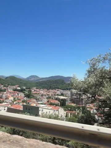 The video pans across a coastal city under a clear blue sky. The foreground features a metal railing and some greenery. In the mid-ground, buildings with red-tiled roofs and modern structures are visible. The background showcases a tree-covered hill sloping down to the sea. The ocean is a deep blue, with a small island in the distance. The camera movement suggests a view from a moving vehicle or a stable elevated platform.