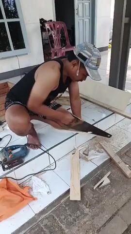 A man wearing a black tank top, patterned shorts, and a baseball cap is crouching on a tiled floor outdoors. He is using a hand saw to cut a piece of wood. Next to him on the floor is a blue jigsaw and an orange cloth. The background shows a building with a door and windows.