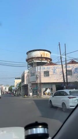 A vertical video shows a water tower built on top of a light-colored commercial building. The water tower is rusted and appears old. The building has multiple floors and windows. The lower part of the building has large, rolled-up metal doors, suggesting it might be a store or garage. There is a street in front of the building with some traffic. A white car is visible on the right side of the road. On the left side, a motorcycle rider is passing by. The sky is clear and blue, indicating it is daytime. The video appears to be taken from inside a vehicle, possibly a car, as the bottom of the frame shows parts of the dashboard and a black object. The overall scene suggests an urban or semi-urban environment.