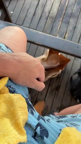 The video begins with a close-up of a person's hand and legs wearing swim trunks. A yellow towel is draped over their legs. A brown paper bag with a clear plastic cup containing a beverage is visible behind their hand. The scene transitions to a wider shot of a swimming pool area. People are seen swimming in the pool, lounging on inflatable chairs, and sitting under umbrellas on a wooden deck. The pool is blue and reflects the sky, which is partly cloudy. There are some trees and buildings visible in the background. The focus shifts to a white plastic bag in the foreground, partially obscuring the view of the pool area.
