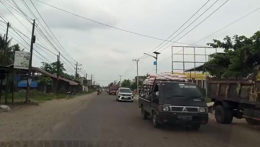 This video captures a perspective from a moving vehicle on a moderately busy road in Keudeteupinpunti, Indonesia. The scene is characterized by a mix of urban and semi-rural elements. On the left side of the road, there are several simple, weathered structures with corrugated metal roofs, some appearing to be shops or small businesses, with motorcycles parked in front of them. Further down the road on the left, palm trees are visible, indicating a tropical environment.

The road itself is paved and appears to be a main thoroughfare, with several vehicles, including a red bus, a white MPV (likely a Toyota Avanza based on the visible front grill), and other cars and motorcycles, traveling in the same direction. The activity taking place is general road traffic and movement within the town.

On the right side of the road, there are more substantial buildings, including a yellow multi-story structure and other constructions, some of which appear to be undergoing development, indicated by scaffolding and building materials. A large billboard structure is also visible on the right.

The sky is overcast with scattered clouds, suggesting it might be late morning or afternoon, and the weather appears to be neutral, possibly humid as is typical for the region. The overall atmosphere is that of everyday life and commerce in a developing area. Overhead, a complex network of power lines crisscrosses the sky, a common sight in many parts of Indonesia. No discernible emotions are conveyed, as the footage is objective and observational. There is no visible text that can be clearly read within the timeframe of the video.