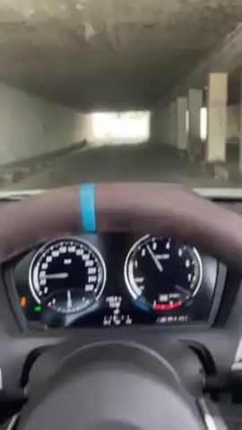 From the driver's perspective, a car accelerates from a tunnel to the outside. The steering wheel, with a blue stripe, is visible in the foreground, along with the car's dashboard. The car is in M Sport mode. The tunnel has concrete walls and support pillars. Outside, there is a clear sky and residential area with houses and trees.