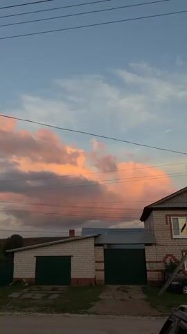 A vertical video captures an outdoor scene with a sky filled with pink and orange clouds. In the foreground, there are two garages with green doors. To the right of the garages, a two-story house is visible, constructed with brick. Several power lines crisscross the sky.