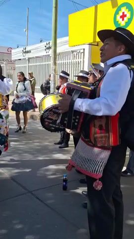 A group of people in traditional clothing are dancing in the street. The dancers are wearing colorful outfits with hats and skirts. Some of the dancers are playing musical instruments, including an accordion and a drum. The street appears to be in a town or city, with buildings and a gas station visible in the background. The sun is shining and the sky is blue.