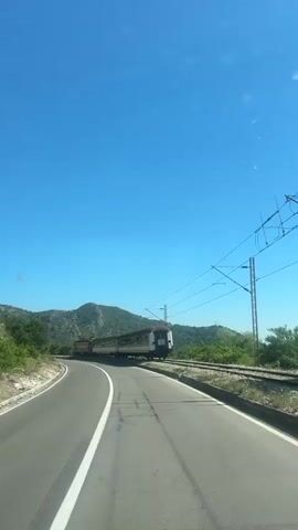 The video, shot from inside a car, shows a train on a track next to a road. The train is composed of multiple passenger cars, painted in shades of green and white, being pulled by an orange and red engine. The road is paved and has a white line marking the edge of the lane. The sky is clear and blue. Low, green mountains are visible in the background. The car is driving forward, parallel to the moving train. Power lines run above the train tracks.