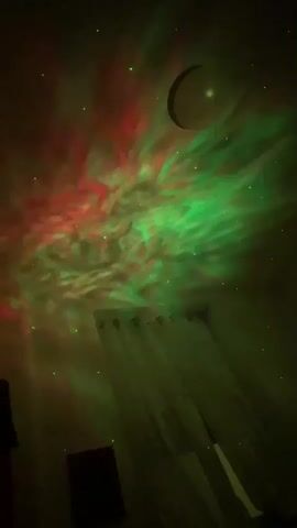 This video captures a shot of a ceiling at night, illuminated by a projector. The projector casts a nebula-like display of red, green, and purple clouds, interspersed with small, green star-like dots. A dark, crescent-shaped object, possibly representing the moon, is visible in the upper portion of the frame. Below the projection, the top of a window with sheer curtains can be seen. The overall impression is that of a projected starry sky or galaxy.