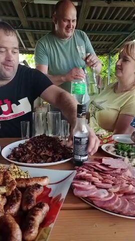 A group of people are gathered around a wooden table laden with food, appearing to be at an outdoor gathering. A bald man in a green t-shirt is pouring liquid from a green plastic bottle into a glass held by a woman in a yellow t-shirt. To the left, a man in a black t-shirt with a red logo is also present. On the table, there are plates of food including sausages, meat, and vegetables, as well as several glasses. The environment suggests a backyard or patio setting with a roof overhead and greenery in the background.