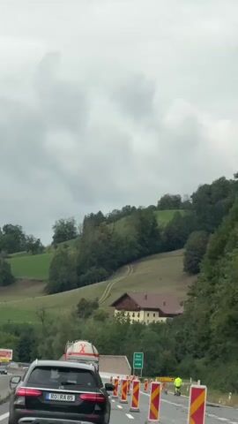 The video shows a view from inside a moving vehicle on a highway. The road has construction barriers and signs. A black SUV is in the foreground, and other vehicles, including a tanker truck, are visible on the highway. The background features a green, hilly landscape with trees and a building with a brown roof. The sky is overcast. A person wearing a high-visibility yellow jacket is seen standing near a sign that says "Ausfahrt", which means "Exit" in German.