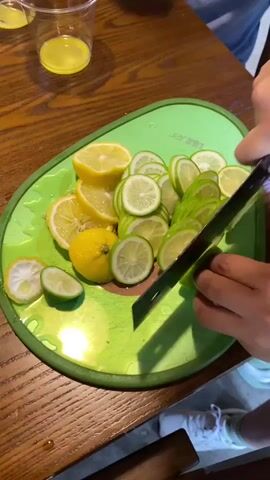 A high-angle, medium shot shows a person slicing limes on a green cutting board. The cutting board is on a wooden table. There are several sliced lemons and limes on the cutting board, as well as a few whole limes and lemons. There are also two clear plastic cups with yellow liquid in them on the table, and a person's legs and feet in white sneakers are visible in the lower right corner of the frame. The person is using a large, dark-colored cleaver with a wooden handle to slice the limes. The scene appears to be indoors, possibly a kitchen or dining area.