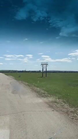 The video shows a view from a vehicle driving on a cracked asphalt road through a grassy, rural area. The sky is blue with scattered white clouds. A sign reading "Капсал" stands to the right of the road. The vehicle's interior elements, such as a grab handle and side mirror reflecting the trailer being towed, are visible.