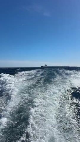 The video shows a wide shot of the ocean from a moving boat, creating a wake of white foam. In the distance, a large gray ship with a helicopter visible on its deck is sailing across the water. The sky is clear and blue, with a hint of land visible on the horizon. The sun appears to be shining brightly.