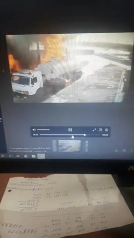 The video shows a computer screen displaying a video of a white tanker truck engulfed in flames. Black smoke is billowing from the front of the truck.  Below the screen is a desk with a piece of paper covered in handwritten numbers and notes.  The video appears to be paused.