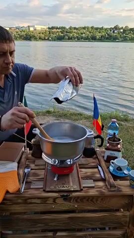 A man is outdoors by a body of water, standing behind a wooden table. He is wearing a dark blue polo shirt. He is holding a silver bag in his right hand and a wooden spoon in his left hand, which he is using to stir a pot sitting on a red portable stove. The pot appears to contain a liquid and some solid objects. The man is looking down at the pot and the bag he is holding. On the table, there are several items, including what appears to be a book, a coffee grinder, a coffee cup and saucer, and a bottle of water. Two small flags, one green, red, and yellow, and another with blue, yellow, and red stripes, are planted in the ground behind the table. The water has trees and buildings on the opposite shore. The sky is cloudy and the sun is setting.