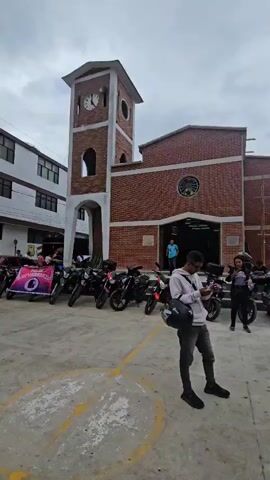 A person wearing a light pink hoodie and dark grey pants stands holding a black helmet and looking at their phone in the foreground of an outdoor scene. The background features a brick church with a clock tower and numerous motorcycles parked in front of it. Several other people are visible near the motorcycles. The ground is paved, and the sky is overcast.