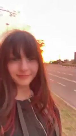 A person with dark hair and bangs, wearing a black top, is holding a phone camera up and recording themself at eye-level. They are standing next to a curb and a multi-lane road. Behind the road are various buildings and trees in the distance. The sky is orange and yellow, indicating it is either a sunrise or sunset. The person speaks into the camera, then opens their mouth wide and appears to scream.