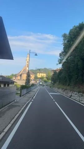 The video shows a road in a small town. On the left side of the road, there are houses and a church with a bell tower. Trees are on the right side of the road. The sky is blue with a few clouds. The road is paved and has white lines marking the lanes. There are a few vehicles visible, including cars and vans. The video appears to be shot from a moving vehicle.