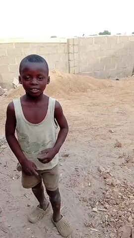 A young boy stands in a dirt field. He wears a white tank top with a light green design, brown shorts, and brown slip-on shoes. He is looking at the camera and smiling, and moves his hands and shifts his weight from foot to foot. Behind him is a cinderblock wall and a large pile of sand. The ground around him is dirt, with small rocks and pieces of debris. The lighting is bright and natural, suggesting it is daytime.