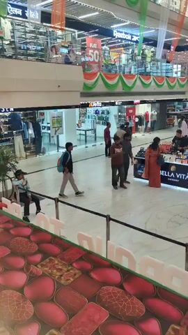 The video pans across a multi-level indoor shopping mall decorated with the Indian flag colors. People are seen walking and browsing in the central atrium area. Several shops are visible on different levels, with signage indicating sales and brands like Reebok, Bella Vita, and Sugar. A kiosk selling cosmetics is prominent in the center. The floor is a light-colored marble, and there are decorative elements at the lower foreground that appear to be a colorful carpet or flooring design. Escalators and railings are also visible, indicating the mall's structure. The overall atmosphere suggests a busy but orderly commercial environment.