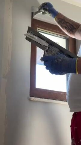 The video shows a person actively plastering a wall around a window frame. The individual is wearing a white t-shirt, red pants (partially visible), and blue work gloves. Tattoos are visible on their right forearm. In their left hand, they hold a large metal trowel, partially loaded with a light gray, pasty substance, likely plaster or putty. A brand name "PRO" is faintly visible on this trowel. With their right hand, they use a smaller, angled plastering tool to apply the substance from the larger trowel to the wall.

The activity focuses on the top-left corner of a brown-framed window. The wall itself is mostly light gray, with an un-plastered section visible on the far left of the frame, revealing a darker underlying surface. Bright daylight streams through the window, indicating it is daytime with clear weather conditions outside. The scene takes place indoors, suggesting a room or building undergoing renovation or construction. Faint text "CE" is visible on the blue glove, likely a certification mark. The individual appears focused on the task of smoothing and finishing the wall surface around the window.