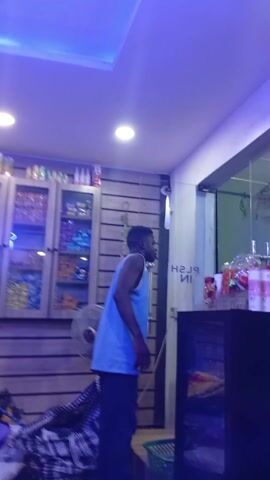 The video captures the interior of a small retail establishment, possibly a convenience store or shop, in Utagban, Nigeria. The space is brightly lit by artificial ceiling lights, with a distinct blue hue cast over the scene, particularly from one of the circular lights.

A young man, dressed in a light blue tank top and dark trousers, stands in the center-right of the frame, facing towards the right. He appears to be observing something out of view or looking towards the exit. His expression is neutral.

To the left, a large display cabinet with glass doors is visible, filled with various packaged goods like snacks and possibly drinks, many in colorful blue and purple packaging. Above this cabinet, additional shelves hold more products, including bottles. The walls are paneled with horizontal wooden slats. A standing fan is positioned on the floor near the display.

At the bottom left, a pile of textiles or clothes, featuring a black and white checkered fabric, is gathered on the floor. On the right side of the frame, a glass door is present, displaying the reversed text "PUSH IN," indicating the viewer's perspective is from inside the shop. Adjacent to the door, there's a counter or shelf holding several items, potentially candies or small goods, and a dark object resembling a monitor or television. A green plastic basket rests on the floor near the counter. The setting suggests a local, small-scale commercial space.