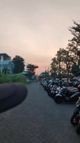 The video shows a paved parking lot filled with motorcycles and several cars parked along the right side of the frame. The motorcycles are lined up neatly, facing forward, with their headlights and mirrors visible. The cars are also parked in a row, extending further down the lot. On the left side of the frame, there is a grassy area with some vegetation and a modern building with white siding. A tree is visible in the middle distance. The sky is a pale orange and pink, suggesting it is either sunrise or sunset. The sun itself is partially visible, low on the horizon, behind a cluster of trees. There are a few people walking in the distance near the parked vehicles. The overall impression is of a quiet, outdoor scene at dusk or dawn.