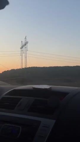 The video shows the view from inside a car driving on a road at dusk or dawn. The sky is a gradient of pale blue to orange. In the distance is a treeline silhouetted against the sky. A power transmission tower is visible on the left. The dashboard of the car, with air vents and a small object on top, is in the foreground. The car appears to be moving.