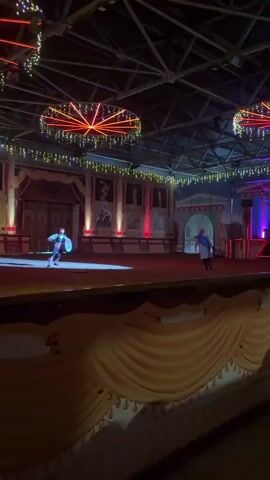 The video shows a dimly lit indoor arena with a stage area. Two individuals, dressed in what appears to be historical or theatrical costumes, are engaged in a mock combat or sword fight. The person on the left wears a green tunic and dark pants with a red sash, holding what looks like two swords. The person on the right wears a blue tunic and white pants, holding a sword and a shield. They are performing in front of a backdrop that resembles an ancient or medieval building facade, with painted murals and architectural details. Festive lights, possibly for a holiday or event, are strung across the ceiling, adding to the atmosphere. The scene is lit by spotlights highlighting the performers, with some ambient lighting in the background. The overall impression is that of a performance or show within a themed venue.
