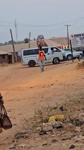 The video shows a dirt road in what appears to be a less developed area. A person in an orange shirt and jeans is walking towards the camera, carrying a large stack of what looks like packaged goods balanced on their head. Another person, wearing a patterned dress and carrying a purse, is walking on the left side of the frame in the same direction.  In the background, there's a white van, a silver car, and a sign that reads "PHL PISHON HOTEL LIMITED". There is sparse vegetation and some litter on the ground. Overhead power lines are visible in the sky.