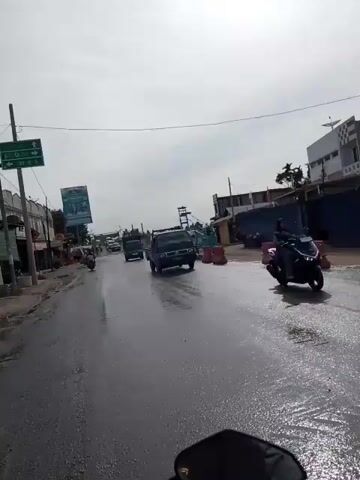 The video shows a street scene from the perspective of someone riding a motorcycle. The road is wet, indicating it may have rained recently or there is some form of water on the surface. Several vehicles are on the road, including a few pickup trucks and cars. On the left side of the road, there are buildings and a billboard. On the right side, there are orange traffic cones and what appears to be construction activity, possibly involving a bridge or overpass. The sky is overcast.