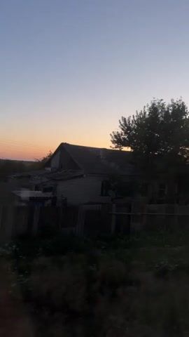 The video captures a rural scene at dusk. A weathered, single-story house with a gray, corrugated roof is visible, partially obscured by trees and vegetation. The sky is a gradient of pale blue transitioning to a soft orange glow near the horizon. A fence runs in front of the house, and in the foreground, there is dense foliage. The overall impression is one of quiet and solitude in a countryside setting.