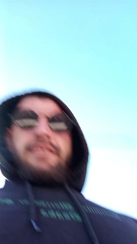 Two men are standing outside. The man closest to the camera is wearing a black hoodie and round sunglasses. He has a beard and smiles at the camera. The man further away is wearing a green jacket. He smokes a cigarette. There is a white van and a house with a red roof in the background. The sky is blue.