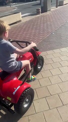 A young boy is riding a red battery-powered three-wheeled toy motorcycle along a brick-paved sidewalk. The boy is wearing a gray shirt and dark shorts. The toy motorcycle has "MIKE" in white letters on the back. He rides in a straight line away from the camera, on a sunny day. In the background, people are walking along the sidewalk, and there are buildings on the other side of the street. The scene appears to be a promenade by the sea.