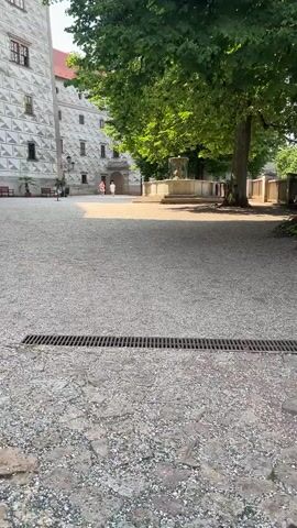 The video captures an outdoor courtyard scene, likely at a historical site or castle. The foreground features a gravel-covered ground with a drainage grate running horizontally across the bottom of the frame. To the left, a section of a large building with distinctive white and grey patterned stonework is visible. The building has multiple windows and a red-tiled roof. In the middle ground, a fountain with a stone basin and a central structure is situated under the shade of a large, leafy green tree. Two individuals, appearing to be women in light-colored dresses, are walking in the distance towards an arched entryway in the patterned building. The lighting suggests it is a sunny day, with distinct shadows cast across the courtyard. The overall impression is one of a tranquil, historical setting.