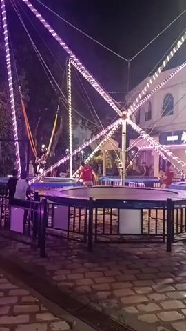 The video shows a bungee trampoline attraction at night, illuminated by purple and white lights. A person is suspended in the air by elastic cords attached to a central structure, having just completed a jump. Below, a trampoline is surrounded by a black fence, with people waiting to participate. On the trampoline itself, another person in a red shirt appears to be assisting the jumper, while other individuals are seated or standing near the attraction. The environment includes paved ground, trees, and buildings in the background, suggesting an outdoor entertainment area.