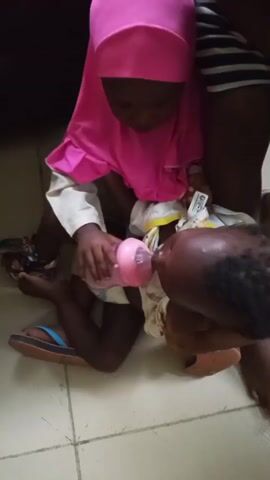 A young child wearing a pink head covering and a light-colored garment kneels on a tiled floor, holding a pink baby bottle. The child is feeding another smaller child who is lying on their back, wearing a white patterned diaper and a light-colored top. The smaller child's head, with dark hair, is visible. An adult's leg or arm is partially visible on the right side of the frame.