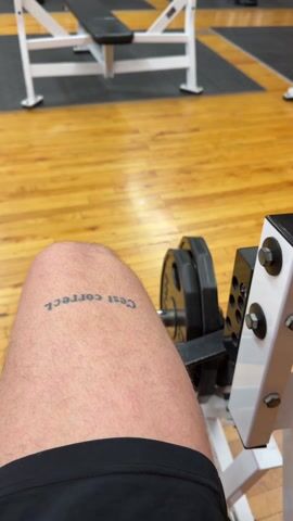 The video shows a person doing leg extensions in a gym. The person is sitting on a leg extension machine with their lower legs hooked under a padded bar that is attached to weights. The person is extending their legs to lift the weights. The person has a tattoo on their upper thigh that reads "C'est correct." The gym has wooden floors, brick walls, and various weight-lifting machines.