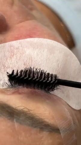 The video shows a close-up of a person's eye during a lash enhancement procedure. A black mascara wand with stiff bristles is being used to comb through the person's eyelashes, which appear to be very dark and full. White under-eye pads are in place to protect the skin from any product. The person's skin is visible around the eye, showing some natural texture and possibly a slight redness or discoloration. The lighting is bright, highlighting the details of the eyelashes and the tool. The background is blurred, suggesting a professional setting like a salon or spa.