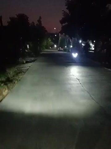 This is a first-person perspective video taken at night while moving down a paved road lined with trees and houses. A motorcycle with its headlight on is visible in the distance, moving in the same direction. Streetlights illuminate the scene intermittently. The sky is dark with a hint of purple or pink near the horizon.