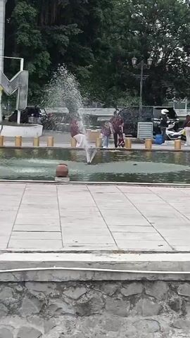 A fountain sprays water into a pool. People stand around the pool. A child is being pushed in a stroller. Trees are in the background.