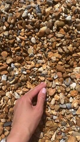 A hand with light pink painted fingernails runs across the surface of small, rounded stones on a beach. The stones are various shades of brown, tan, and white, with some gray stones also visible. The hand is fair-skinned. Sunlight illuminates the scene.