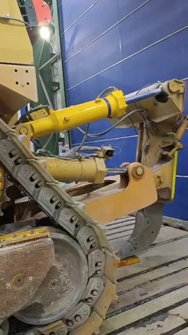 This is a close-up, slightly shaky, vertical video taken indoors, likely in a workshop or garage. The primary subject is the lower half of a large, yellow bulldozer. The video pans slowly to the right, showcasing the heavy-duty track and its components.

In the foreground, the thick, segmented metal track is clearly visible, showing signs of wear and dirt. A large, circular sprocket, also yellow and worn, is prominently featured, with bolts around its circumference. To the left of the sprocket, a part of the bulldozer's undercarriage is visible, appearing rusty and dirty.

Moving further into the frame, a yellow hydraulic cylinder is mounted horizontally, attached to other mechanical parts of the bulldozer. This cylinder has blue components at its ends. In the background, parts of the bulldozer's cabin structure and some industrial shelving are visible. A blue, corrugated metal wall forms the backdrop on the right side of the frame. The lighting appears to be artificial, coming from overhead fixtures. A few glimpses of what might be clothing, possibly a white protective suit, are visible near the middle of the frame, suggesting someone might be working on the machine. The overall impression is of a large, operational piece of heavy machinery undergoing maintenance or being stored.