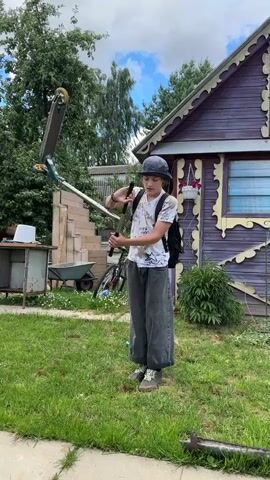 A young person wearing a helmet and backpack is standing on a lawn in front of a purple wooden house. The person is holding onto the handlebars of a scooter. The scooter is green and silver with yellow wheels. The person's left foot is on the ground, and their right foot is on the scooter's deck. The person is looking at the camera and appears to be preparing to ride the scooter. The background shows trees, a wheelbarrow, and a bicycle. The sky is cloudy.