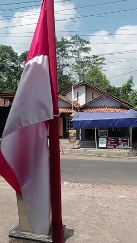 A close-up shot of a red and white flag is in the foreground, blowing in the wind. Behind the flag, a street is visible with a building on the other side. The building appears to be a small shop with a blue awning. There are trees and power lines in the background. The sky is cloudy. The flag is red on top and white on the bottom, with a gold pole. The flag is tied to the pole with a white string. The street is paved and there is a sidewalk in front of the flag. The shop has a sign with text on it, but it is not readable. There is another smaller flag flying on a pole in front of the shop.
