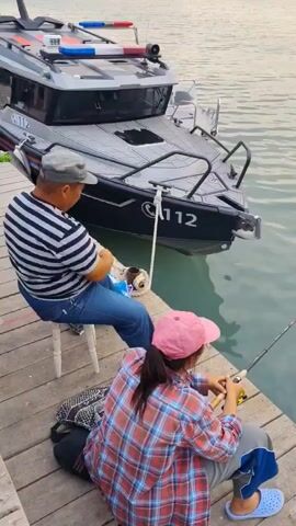 The video shows two people fishing on a wooden dock. A man is sitting on a white stool, wearing a striped shirt and a cap. He is holding a white bag and a rope attached to a boat. A young woman is sitting on the dock in front of him, wearing a plaid shirt and a pink cap. She is holding a fishing rod. Behind them, a black boat with "112" written on it is docked. The water is calm, and the sky appears overcast.