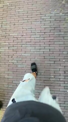 A first-person perspective video shows a person walking on a brick-paved sidewalk on a sunny day. The person is wearing light-colored ripped shorts and dark slides. To the left, a silver car is parked parallel to the street. The street is lined with brick buildings, some with potted plants in front. The person walks towards a narrow street with more parked cars and then turns to face the street. The camera then pans to show the street continuing with more brick buildings on either side. A flag is visible hanging from one of the buildings.